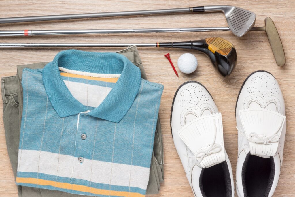 Golf apparel and equipment laid out on a wooden surface, including a sustainable polo shirt, a pair of shoes, clubs, a tee, and a golf ball.