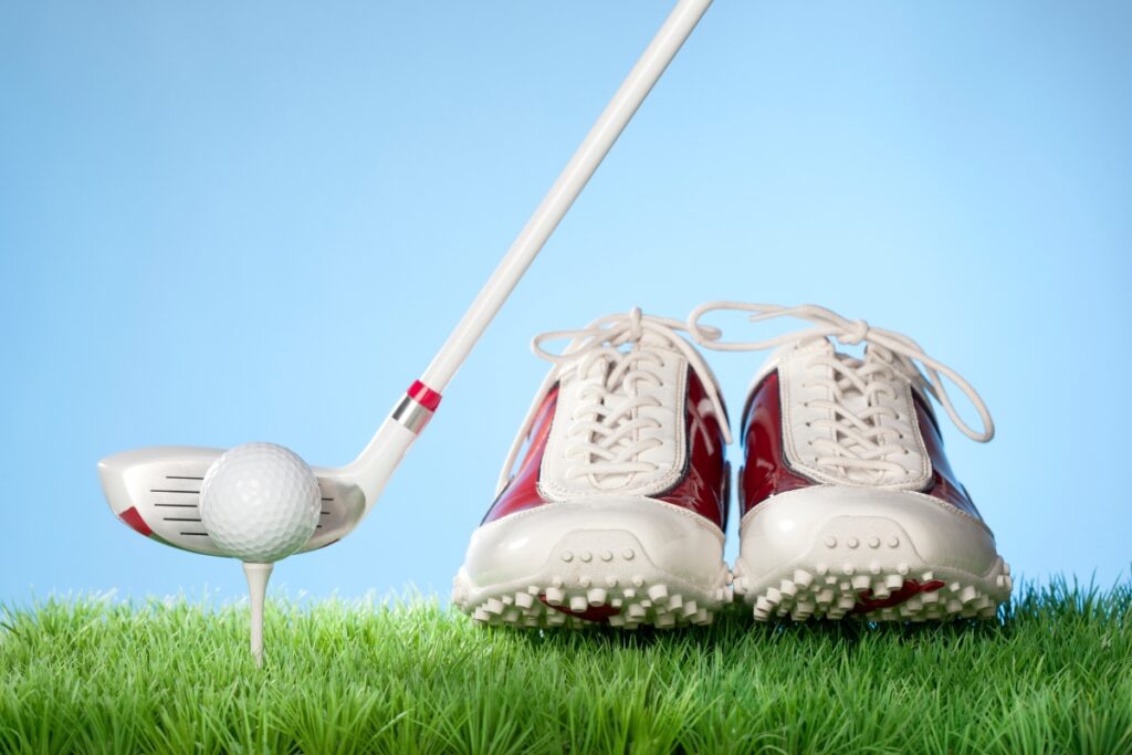 A golf ball on a tee with a club equipped with the latest golf technology and a pair of white and red golf shoes on grass against a blue sky background.