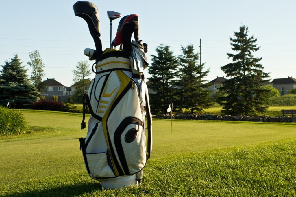 Golf bag with clubs and accessories on a course near a green with a flag pole, trees, and residential homes in the background during daytime.