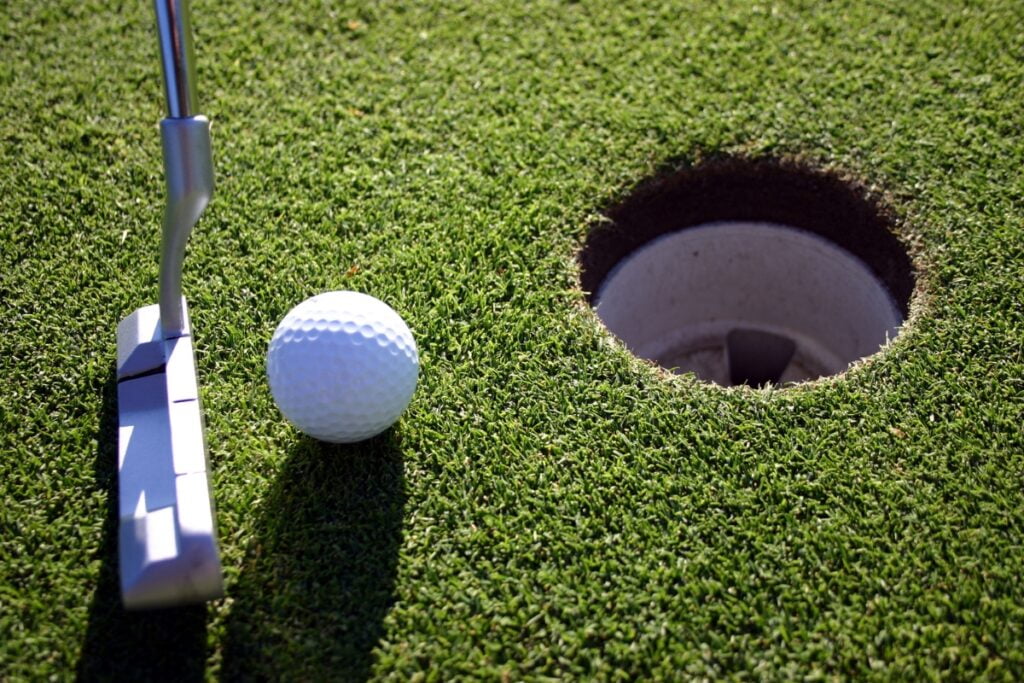 A close-up view of a golf ball near a hole on a green, with various golf accessories, including a putter positioned to the left side of the ball, indicating an imminent putting action.