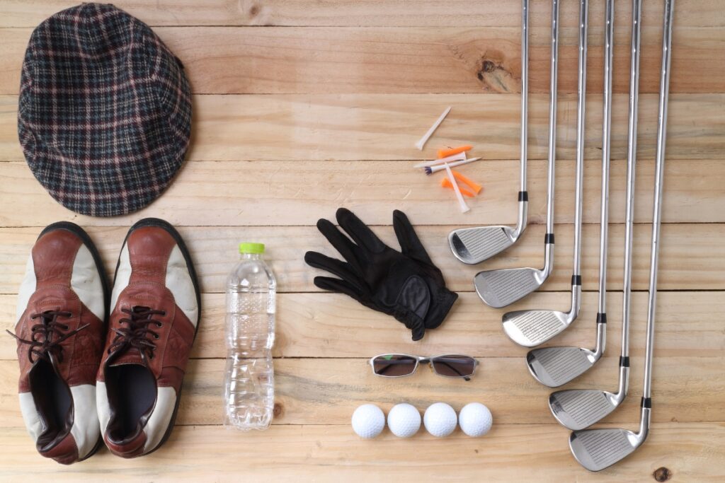 Golf accessories and casual items laid out on a wooden surface, including clubs, balls, tees, gloves, a hat, sunglasses, shoes, and a water bottle.