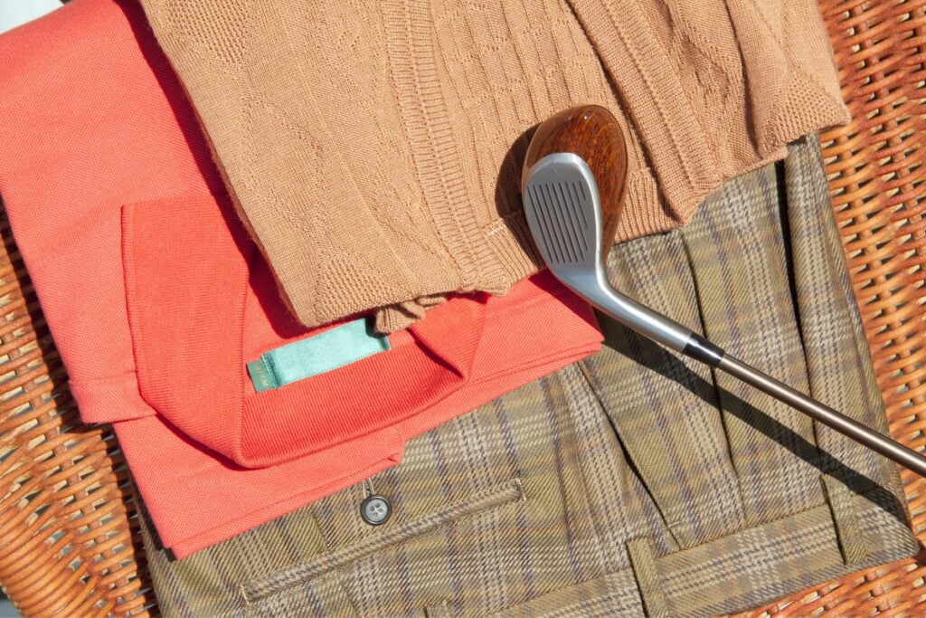 Golf club beside a folded orange sweater from a top golf apparel brand on a plaid patterned picnic blanket in sunlight.