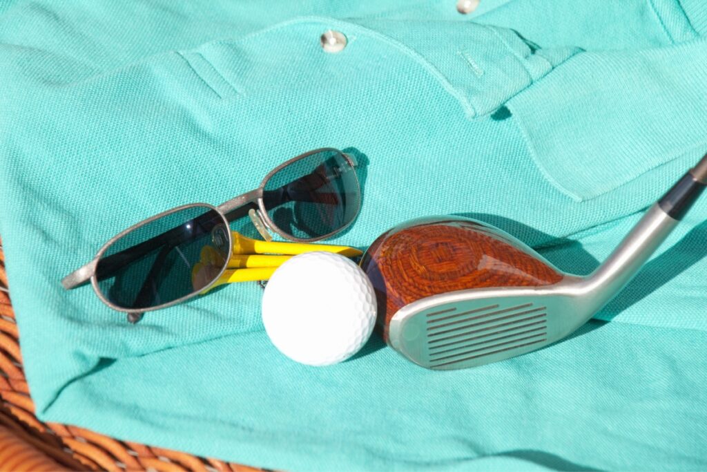 Sunglasses, golf ball, and iron golf club on a turquoise polo shirt from top golf apparel brands, suggesting a leisurely day of golf.