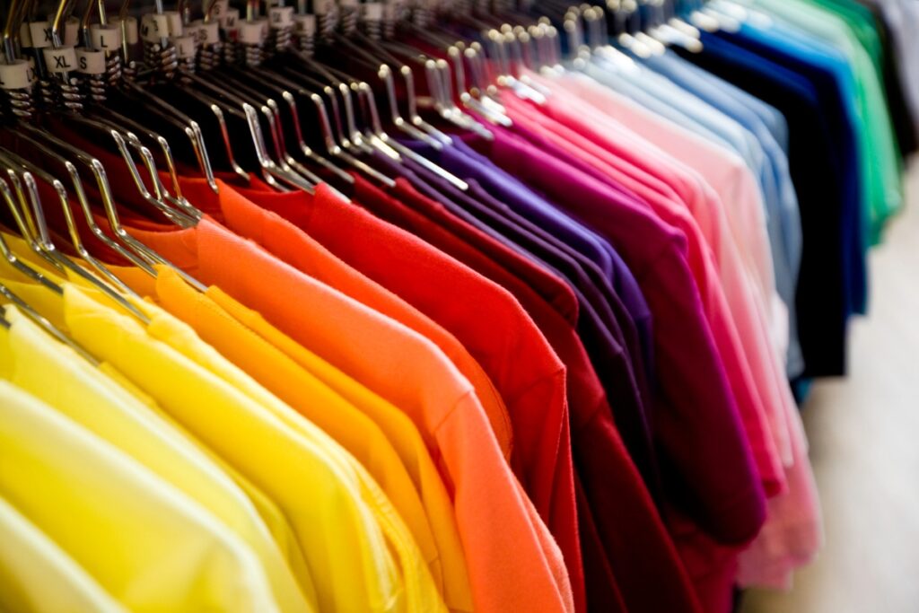 Colorful t-shirts from top golf apparel brands hanging in a retail store arranged in gradient from yellow to blue, with visible size tags on hangers.