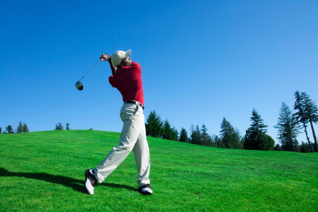 A golfer in a red shirt and white pants, showcasing their golf fitness, swings a golf club on a sunny day, surrounded by green grass and trees.