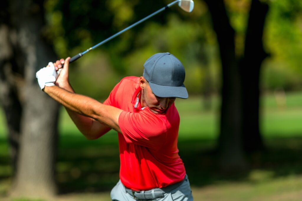 A golfer in a red shirt and gray cap swings a golf club on a green course with trees in the background, showcasing his golf fitness.