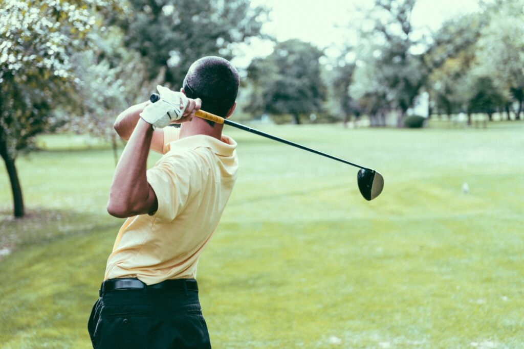 A person in a yellow shirt and black pants, demonstrating impeccable golf fitness, swings a golf club on a lush green course with trees visible in the background.