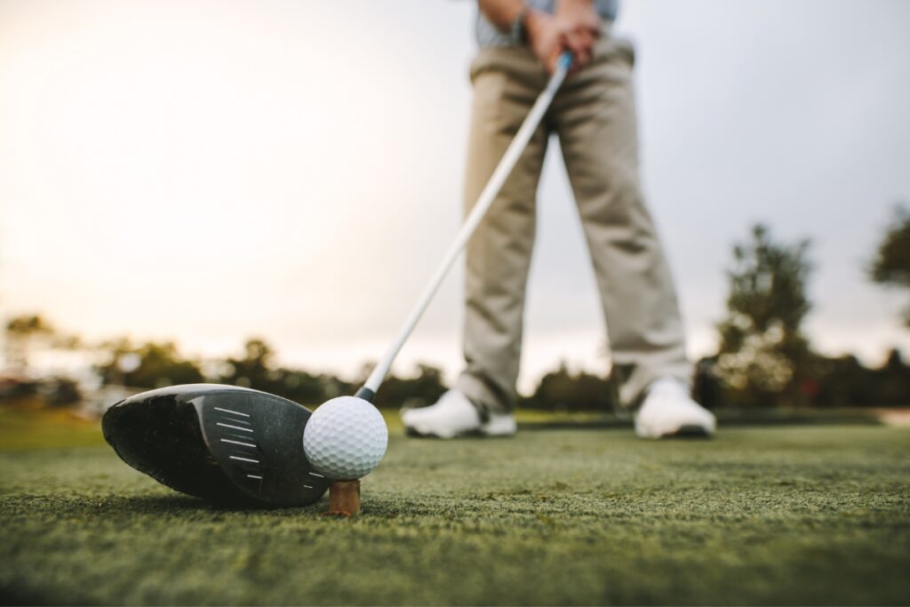 A golfer, showcasing his golf fitness, prepares to hit a golf ball on a tee with a driver club, with the shot taken from ground level focusing on the ball and club.