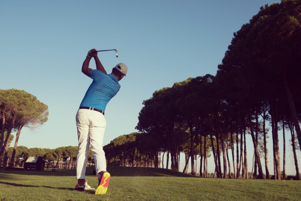 A golfer, seen from behind, follows through on a swing, demonstrating his peak golf fitness as he hits a ball towards a wooded area on a sunny day. He wears a blue shirt, white pants, and a white cap.