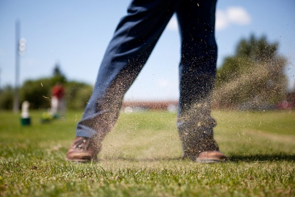 A golfer's legs, exhibiting clear golf fitness, and part of their swing are visible as they kick up a spray of grass and dirt, standing on a vibrant grassy course with trees and a clear sky in the background.