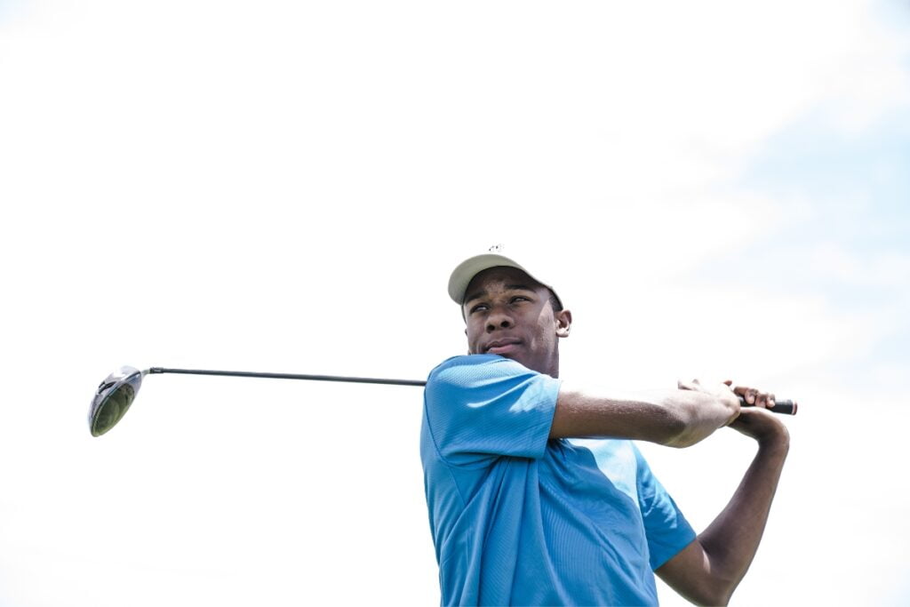 A person wearing a blue shirt and white cap is holding a golf club, appearing to have just swung it. The clear and bright sky in the background emphasizes their commitment to golf fitness.