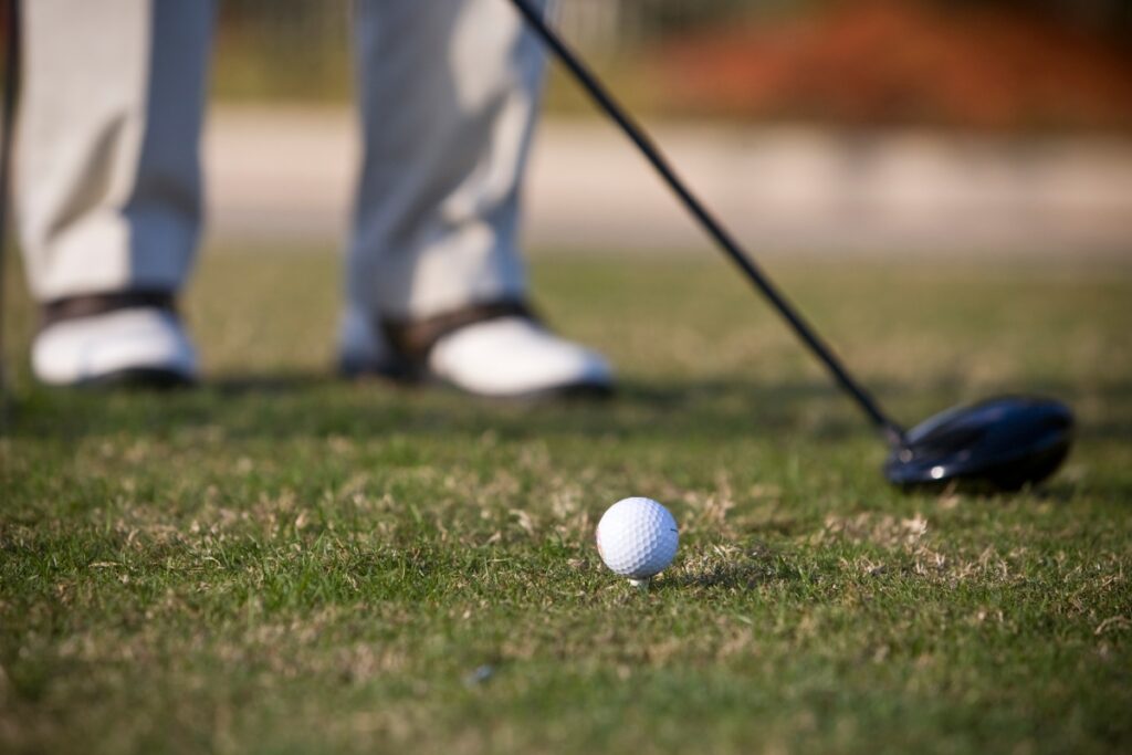 Close-up of a golfer's legs in beige pants and white shoes, standing on grass next to a golf ball and a golf club, embodying peak golf fitness.