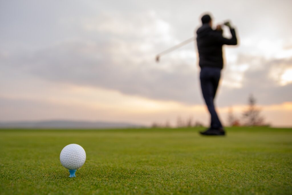 A golfer demonstrates peak golf fitness as they swing a club on a golf course at sunset, with a golf ball on a tee in the foreground.