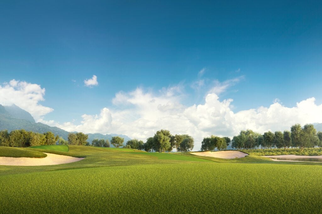 A green golf course with sand bunkers is set against a background of trees, mountains, and a blue sky with scattered clouds, making it one of the most picturesque golf travel destinations.