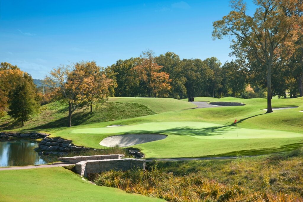 A well-maintained golf course with lush green fairways, multiple sand bunkers, and a small pond under a clear blue sky makes for one of the top golf travel destinations. Trees with autumn foliage are visible in the background.