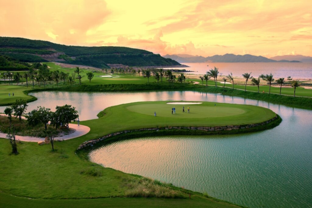 Golfers on a grassy island green surrounded by water at sunset, one of the most picturesque golf travel destinations. Palms and hills stand majestically in the background.