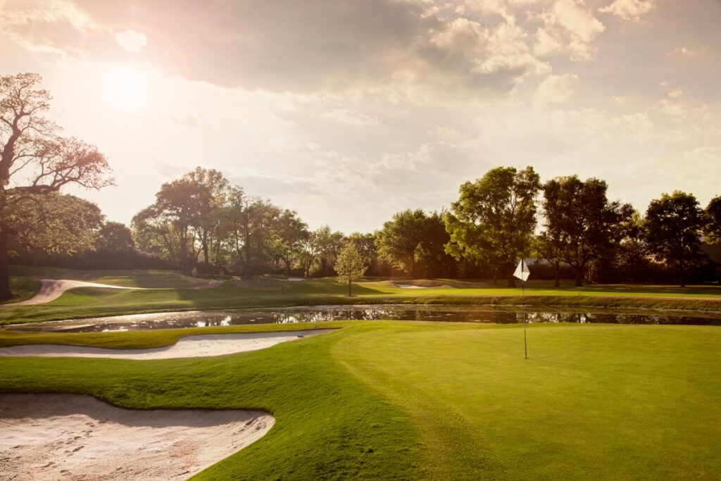 A premier golf travel destination featuring a pristine course with a green, a flagstick, bunkers, and trees in the background under a partly cloudy sky with the sun shining.