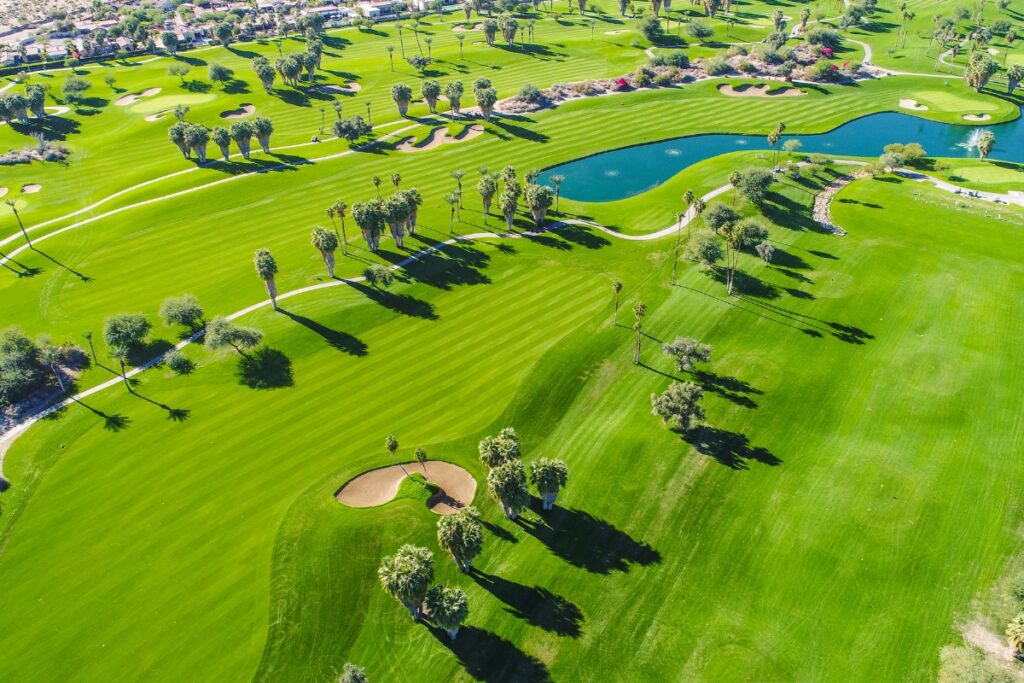 Aerial view of a lush green golf course featuring several sand bunkers, water hazards, and scattered palm trees, with a residential area in the background—an ideal spot for golf travel destinations.