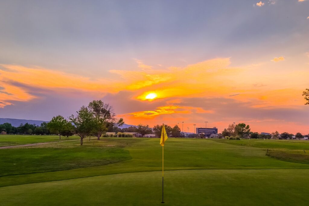 A sunset over a renowned golf travel destination with a green field, a yellow flag marking a hole, trees, and distant buildings.