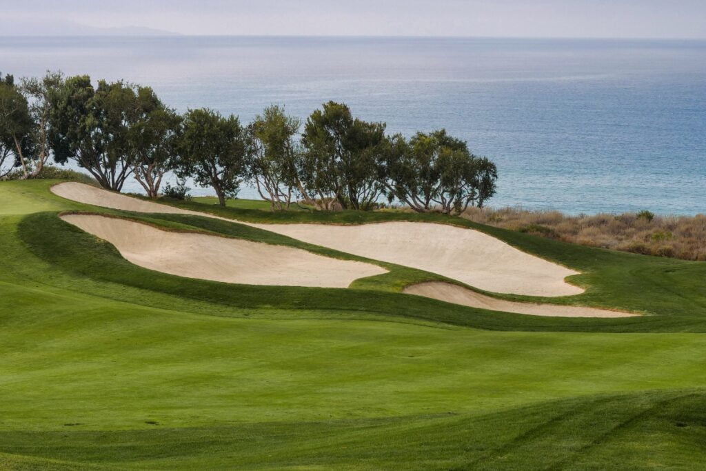 A scenic golf course with sand bunkers, green fairways, and trees is set against an ocean backdrop on a cloudy day, making it one of the premier golf travel destinations.