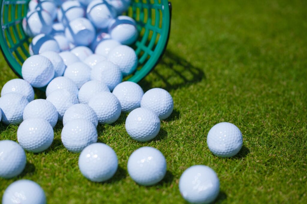 A green basket has been tipped over, spilling numerous white golf balls and a few cutting-edge golf tech gadgets onto a neatly trimmed grass surface.