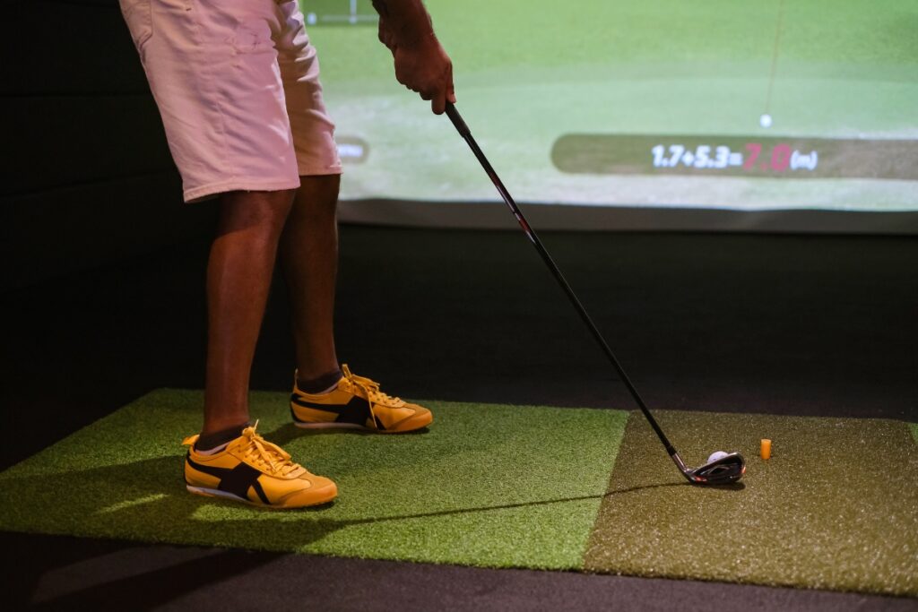 A person in white shorts and yellow shoes prepares to swing a golf club on a patch of artificial turf indoors, surrounded by cutting-edge golf tech gadgets, with a projection screen showing golf data in the background.