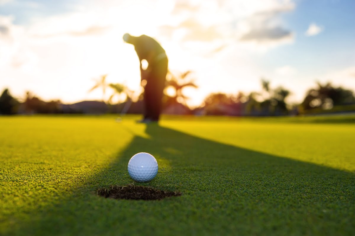 A golfer prepares to putt on a sunny day, with a golf ball near the hole in the foreground, setting a perfect scene for some casual golf business networking.