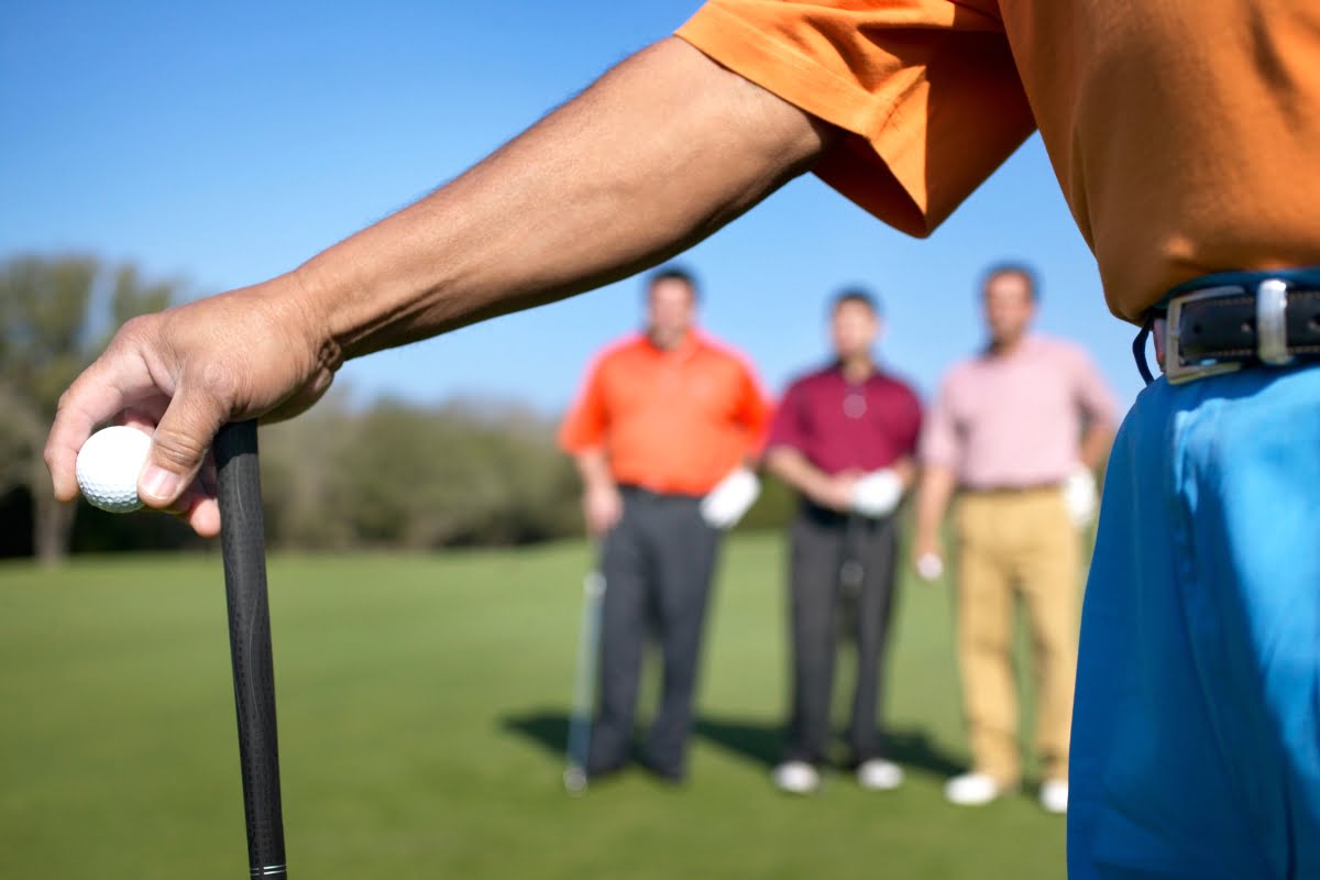 Close-up of a golfer's hand holding a club and a golf ball in the foreground, with three other golfers engaged in golf business networking in the background on a pristine golf course.