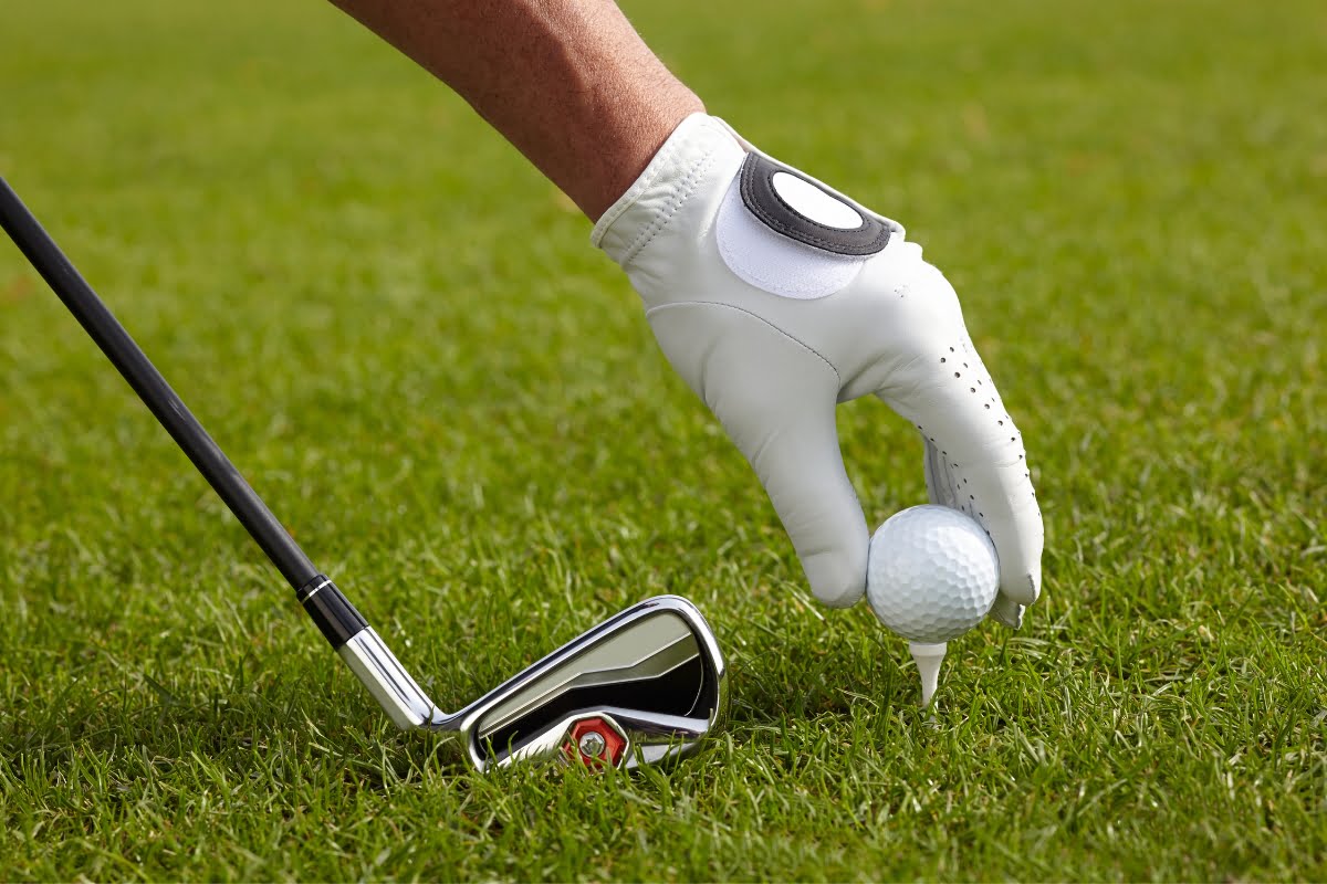 A golfer wearing a white glove places a golf ball on a tee with a golf club positioned next to it on a grassy field, ready to combine the sport with some golf business networking.