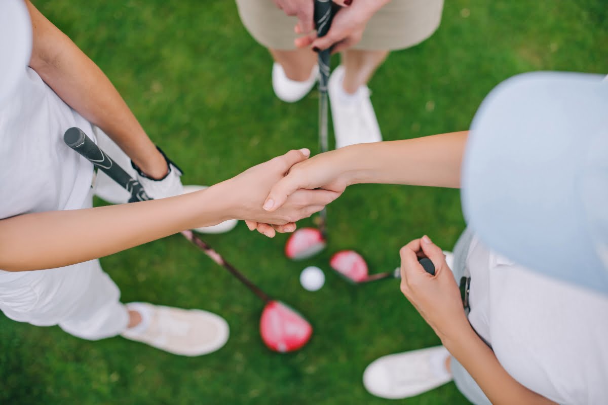 Two individuals shaking hands on a golf course, with golf clubs and a golf ball on the grass, viewed from above. This moment captures the essence of golf business networking at its finest.