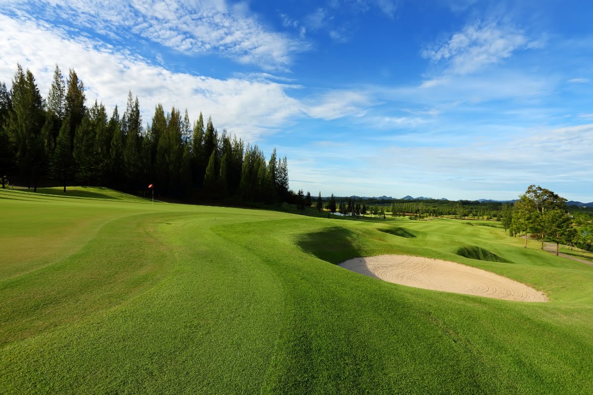 A well-maintained golf course, showcasing expert golf course design, features a green fairway, sand bunker, and a red flag in the distance. Tall trees border the course under a blue sky with scattered clouds.