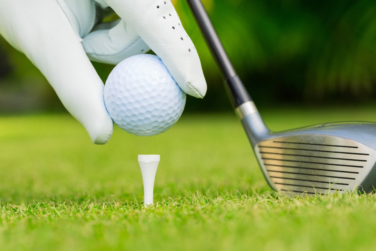 A gloved hand places a golf ball on a tee, with a golf driver club positioned nearby on a meticulously crafted grassy field, showcasing the pinnacle of golf course design.