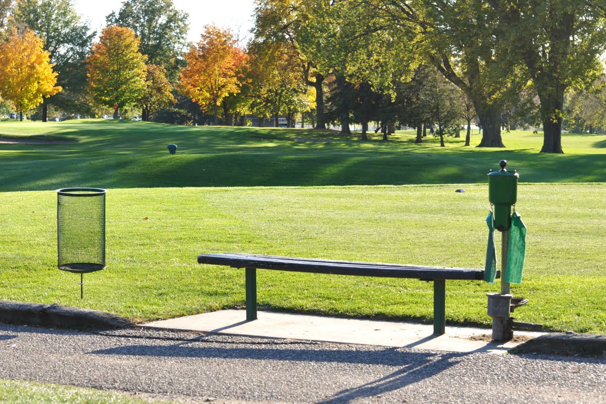 A green park area with a wooden bench, a trash can, and a hand sanitizer station, thoughtfully laid out like elements of a golf course design. Trees with autumn foliage are visible in the background.