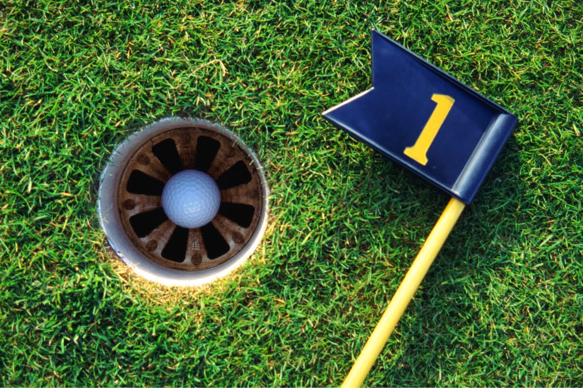 A golf ball resting near the edge of a hole on a meticulously crafted green turf, with a flag marked "1" lying next to it, showcases the excellence in golf course design.