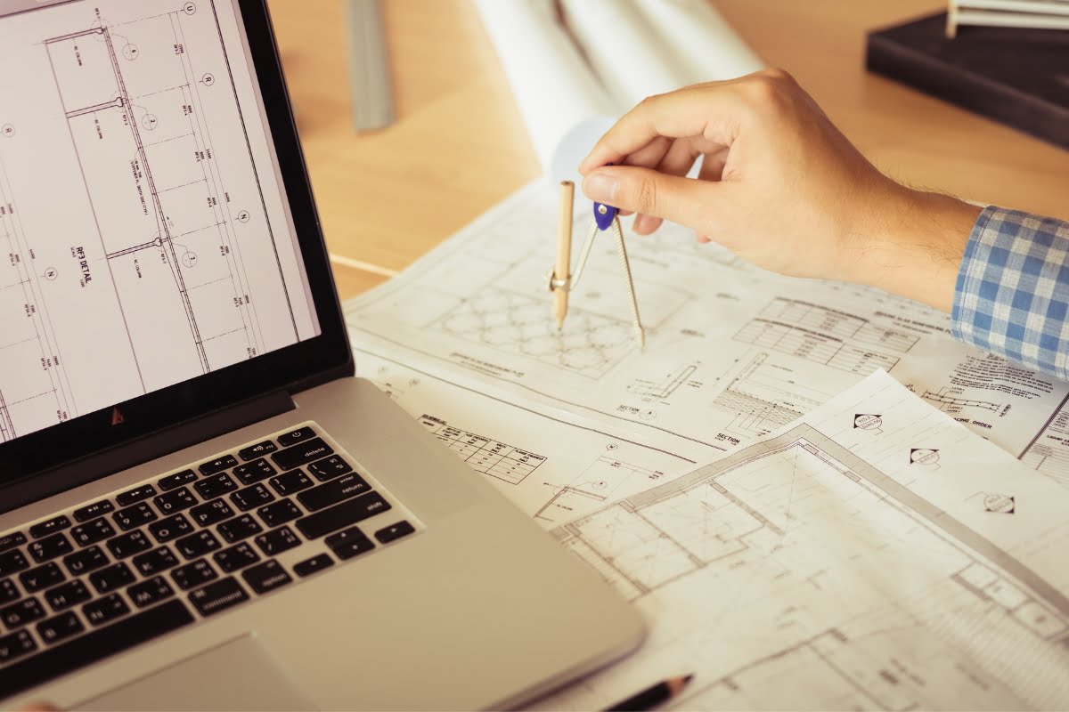 A person in a checkered shirt uses a compass to work on architectural plans for golf course design, spread on a desk next to an open laptop displaying similar plans.