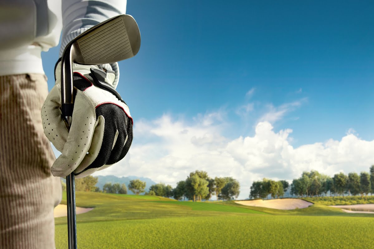 Close-up of a golfer's gloved hand holding a golf club, with the serene golf course and blue sky in the background, embodying a moment of golf mindfulness.