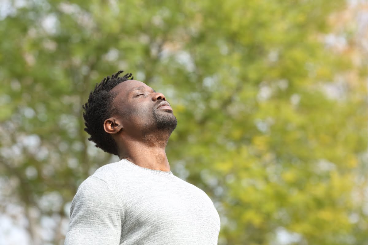 A person with short hair wearing a gray sweater stands outside with eyes closed, head tilted slightly upward, and appears to be taking a deep breath, embodying mindfulness. Green, blurred trees are in the background.