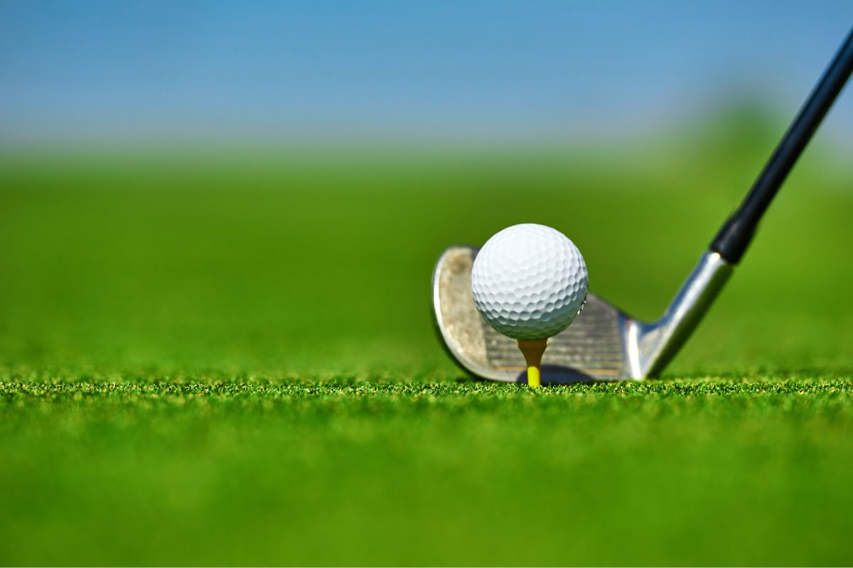 Close-up of a golf club set to hit a ball on a tee, against a serene green grass background with a clear blue sky, capturing the essence of golf mindfulness.