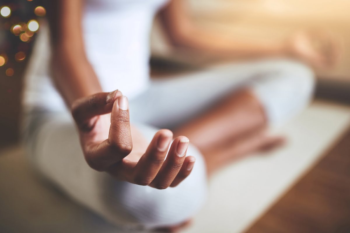 A person sits cross-legged on a mat, meditating with fingers in a mudra position, embodying mindfulness. They wear a white tank top and light gray pants.