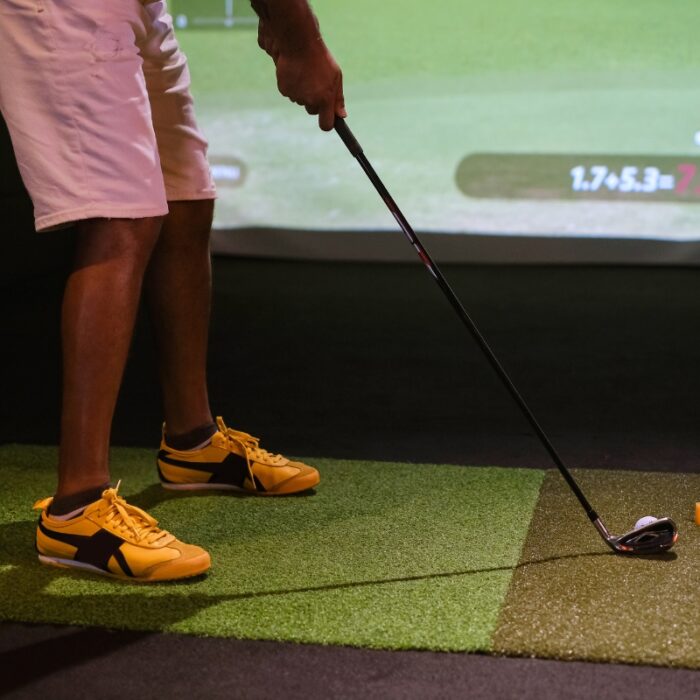 A person lines up a golf shot on an indoor golf simulator, wearing yellow and black sneakers, white shorts, and holding a golf club. The room's stylish golf home decor sets the perfect ambiance for practice.