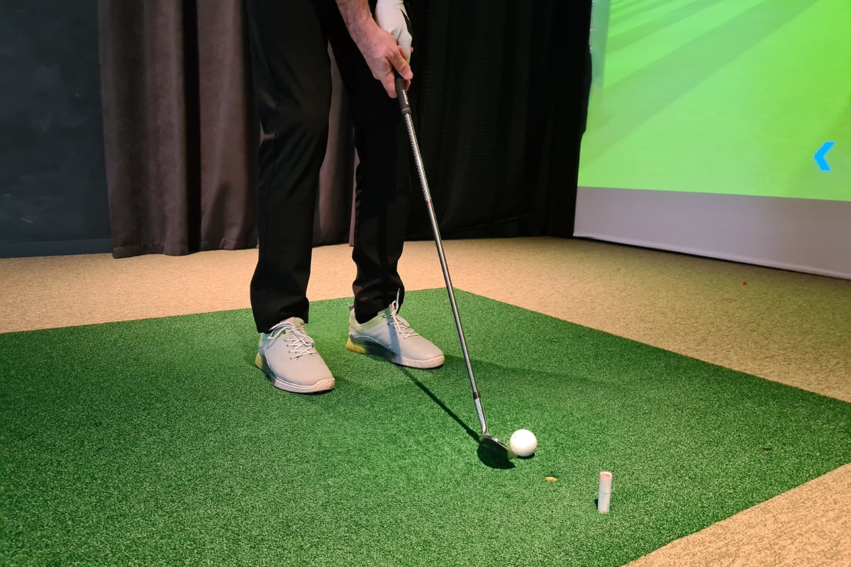 Person in black pants and white shoes holding a golf club, preparing to hit a golf ball on an indoor green turf. A projection screen is visible in the background, adding an element of golf home decor.