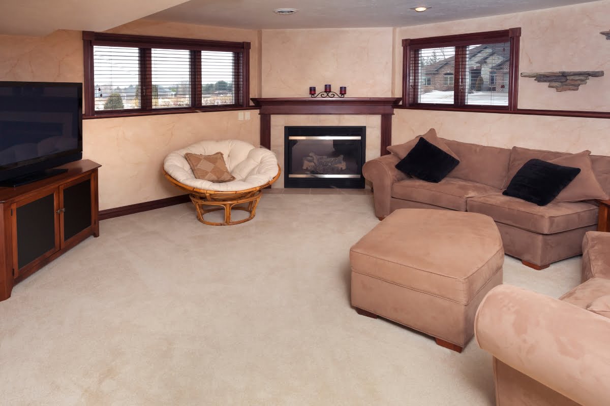 A cozy basement living room features a beige sectional sofa, chair, and ottoman, a wall-mounted TV, and a fireplace with a wooden mantel. The room is carpeted and has two windows with blinds. Subtle golf home decor pieces add a unique touch to the inviting space.