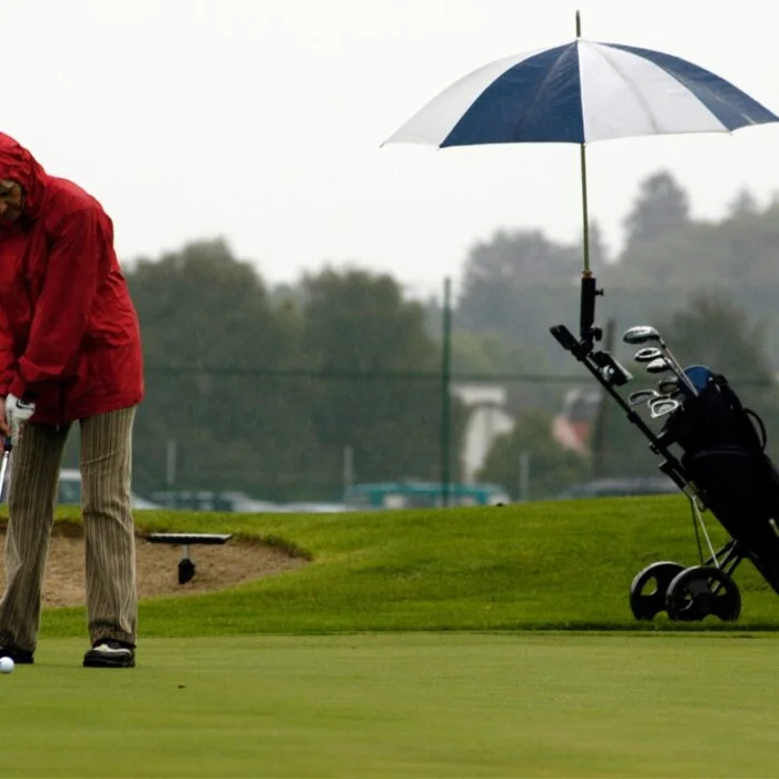 Person in a red rain jacket putting on a golf course while an umbrella covers a golf bag on a pushcart. Despite the gloomy sky and chilly air, they continue golfing in cold weather clothing, determined to enjoy their game. The background features trees shrouded in mist.