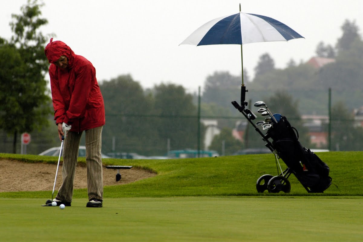 Person in a red rain jacket putting on a golf course while an umbrella covers a golf bag on a pushcart. Despite the gloomy sky and chilly air, they continue golfing in cold weather clothing, determined to enjoy their game. The background features trees shrouded in mist.