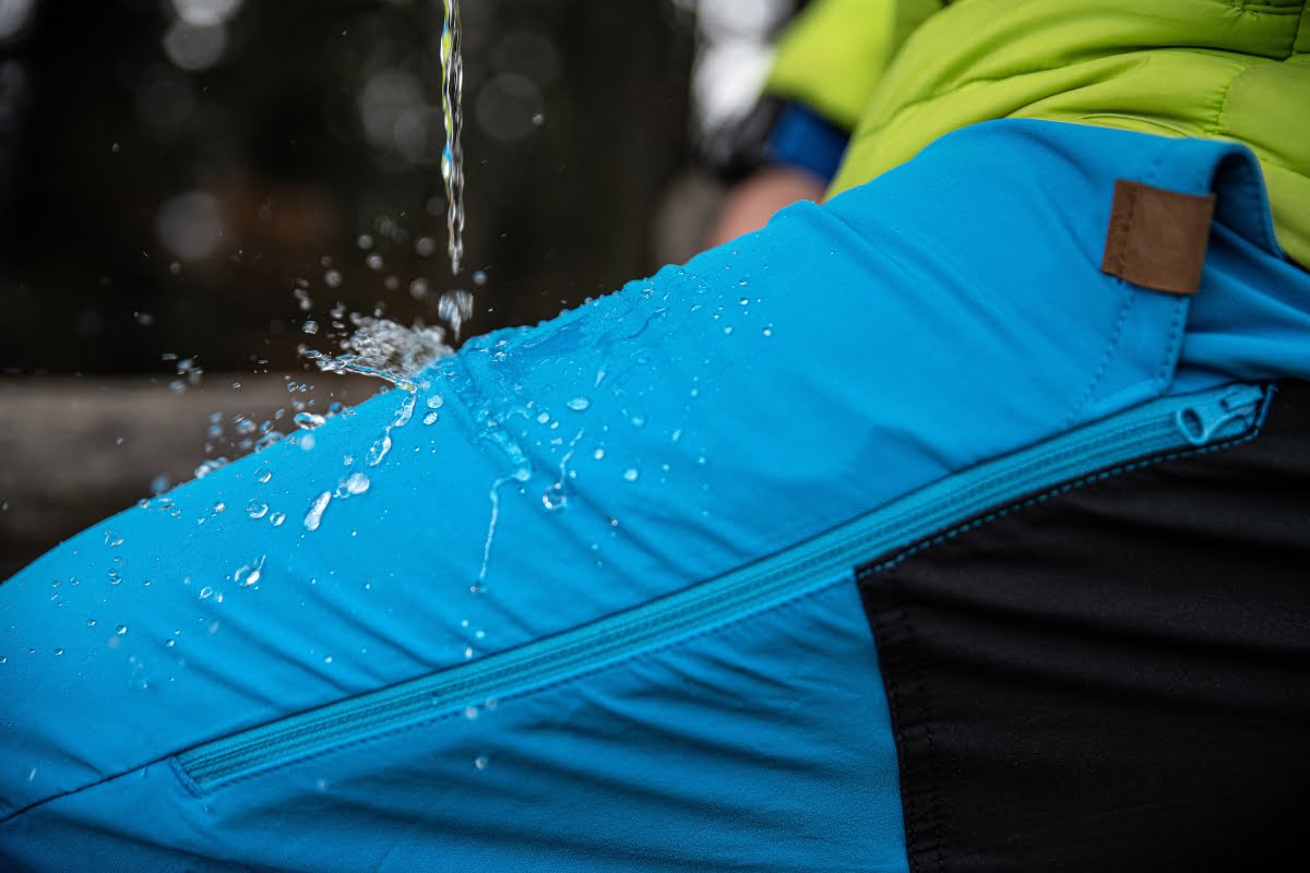 Close-up of water droplets splashing on blue waterproof pants with a zipper, worn by someone in a green jacket, perfect for golfing in cold weather.