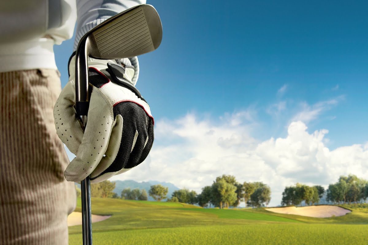 A person wearing a golf glove and cold weather clothing holds a golf club on a green golf course under a blue sky with clouds.