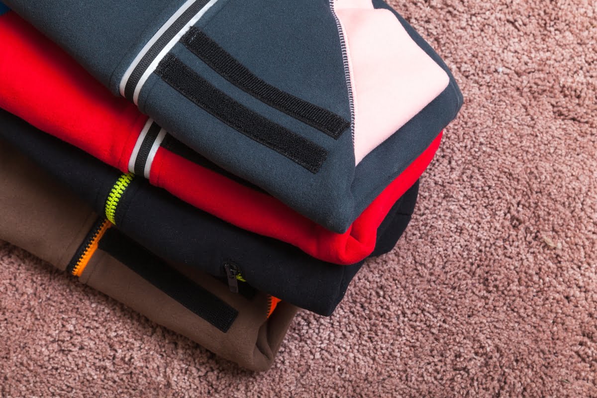 A stack of colorful folded clothing, including red, black, brown, and gray apparel with zippers perfect for golfing in cold weather, rests on a beige carpet.