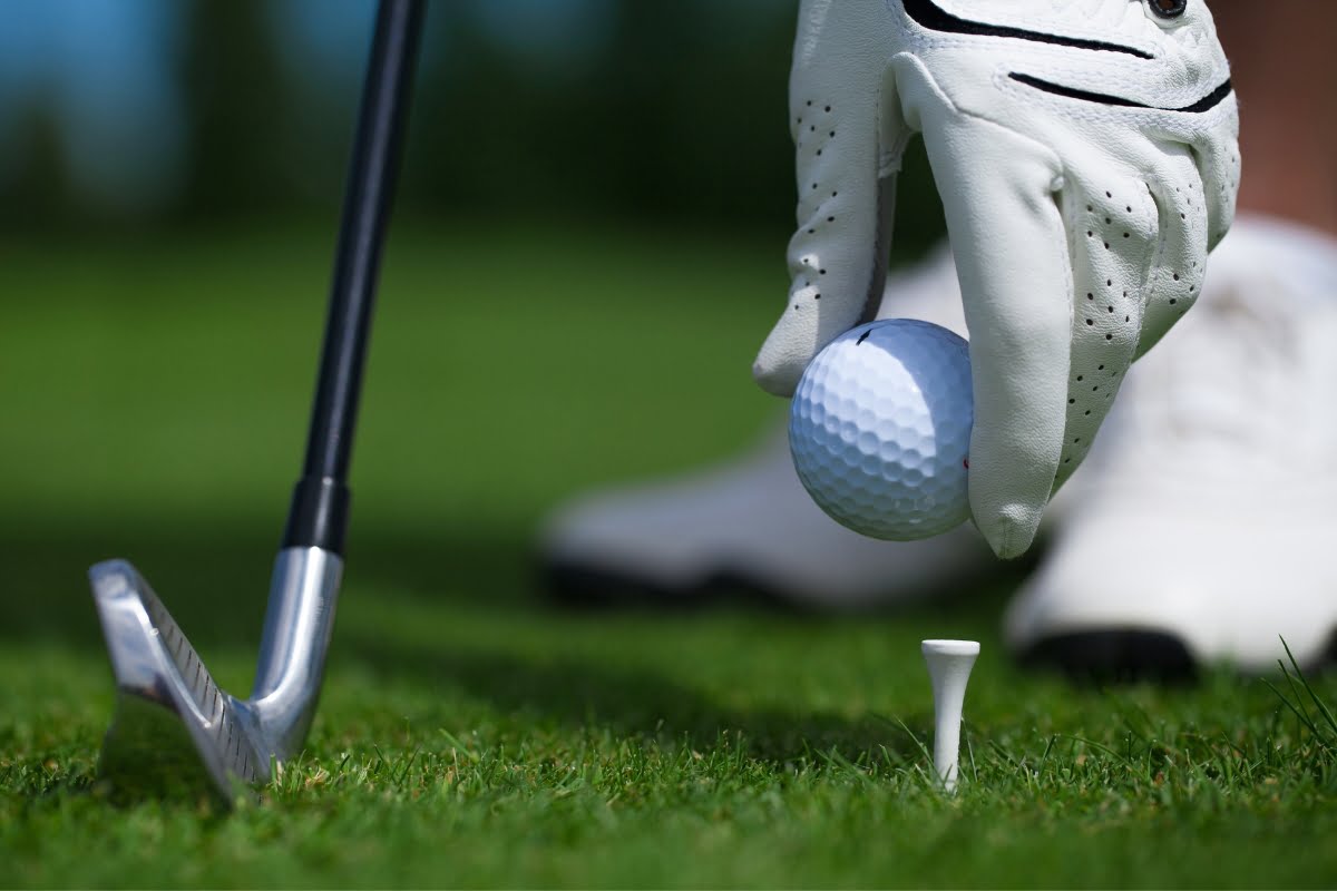 A gloved hand, wrapped in cold weather clothing, places a golf ball on a tee with a golf club positioned nearby on the grassy surface.