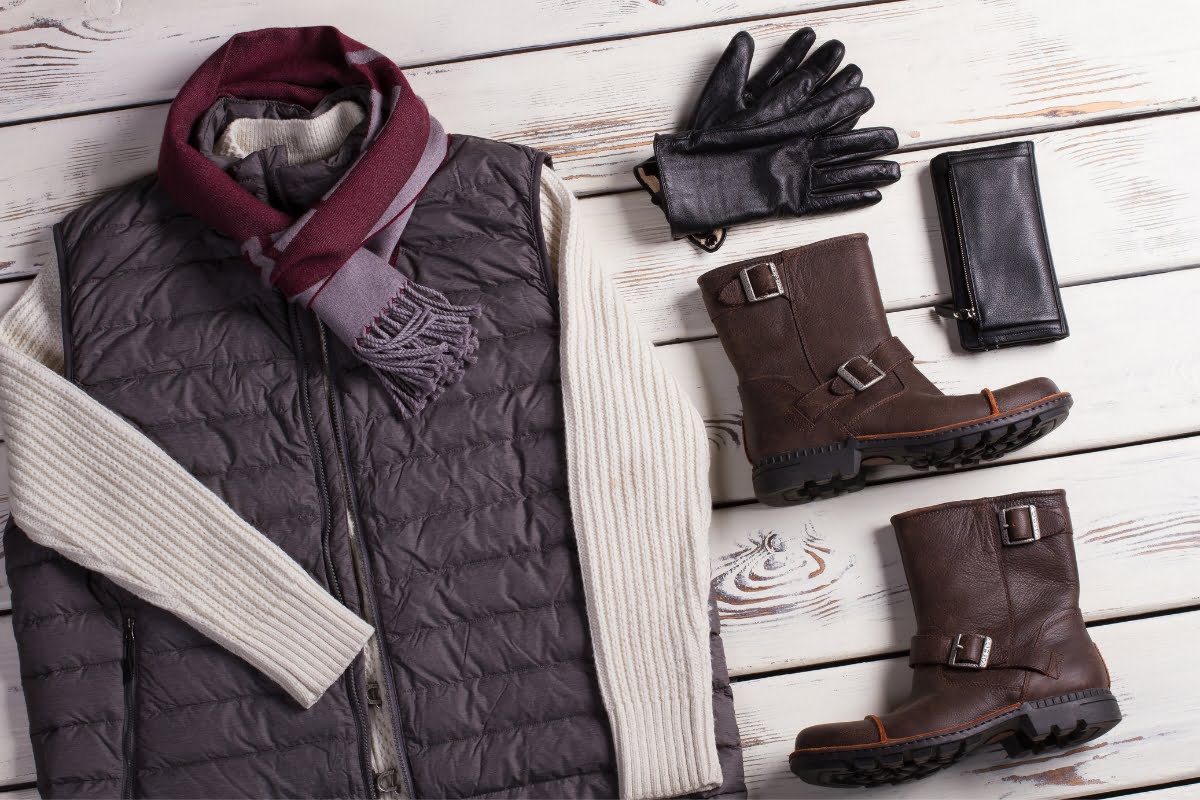 Winter outfit laid out for golfing in cold weather, including a puffer vest, cream sweater, maroon scarf, black leather gloves, brown boots, and a black wallet, all on a wooden surface.