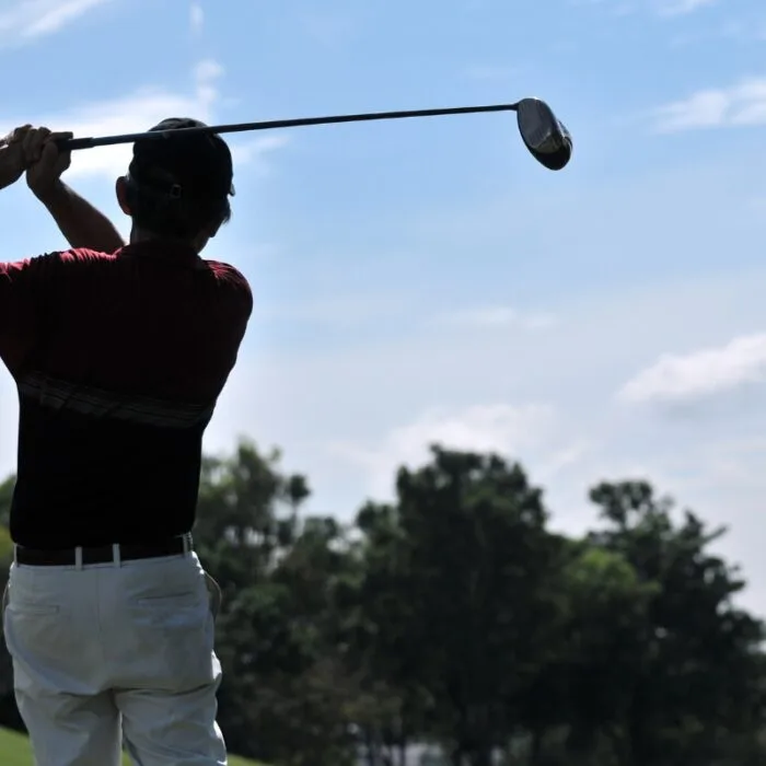 A golfer mid-swing, showcasing impressive golf performance on a sunny day with trees in the background.